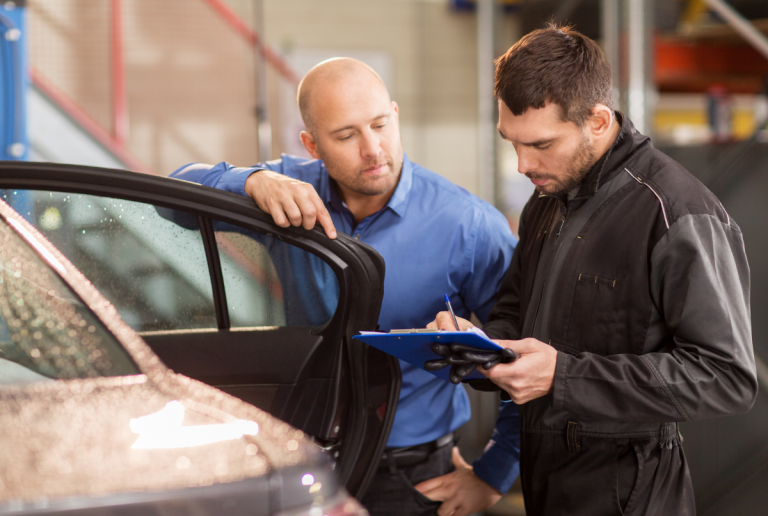 Mechaniker & Kunde stehen am Auto, der Mechaniker zeigt dem Kunden etwas auf seinem Clipboard
